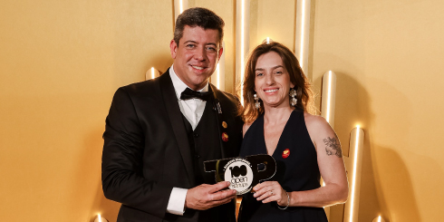Foto de dois representantes em traje formal segurando o troféu do Ranking 100 Open Startups diante de um painel dourado iluminado. A dupla sorri para a câmera durante a premiação.