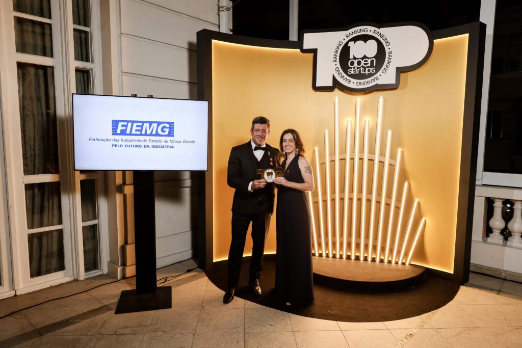 Foto de representantes da FIEMG recebendo um prêmio no evento Ranking 100 Open Startups. A dupla posa diante de um painel iluminado dourado com o logo ‘Top 100 Open Startups’, segurando o troféu. Ao lado, há uma tela exibindo a marca FIEMG. O cenário é elegante e formal, em um ambiente interno.